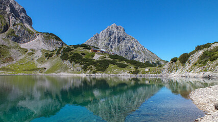 Naklejka na ściany i meble See mit Spiegelung in den Bergen und einer Berghütte bei blauem Himmel