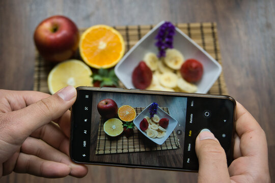 hand of a person grabbing a smartphone or cell phone taking a photo of a dish of food before eating fruit, breakfast and a healthy bowl