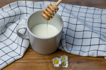 A honey dipper pours honey into a cup of milk on a wooden table with daisies