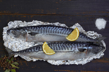 Two atlantic mackerel fishes ready for cooking decorated with spices, salt and lemon slices on the black wooden background. Top view