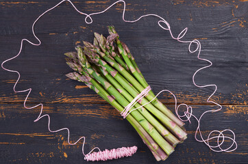 Top view on the fresh green asparagus bunch decorated with pink thread on the black wooden background. Healthy spring vegetables ready for cooking