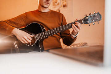 Obraz premium Man learning to play guitar with the help of online learning at home. Guy sitting at table with laptop and black guitar