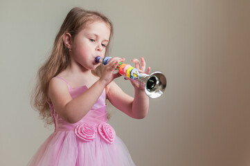 A cute little girl in a lush pink dress plays the toy trumpet.