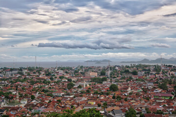 Aerial view of dense city and population Bandar Lampung cityscape with horizon and coastline in background. Strange cloud formation. Cloudy blue sky. 