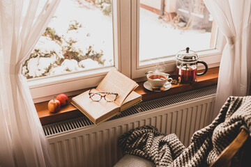 Winter relax at cozy home. Hot tea, teapot and book by the window.