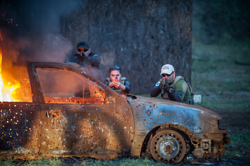 Plakat Army soldier hiding behind burning car shooting from machine gun. Action tactical combat