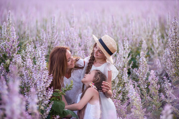 Young mother embraces two daughters among purple blooming sage field. Girls hug together, laugh, have fun. Dressed in white with straw hats. Traveling with children, maternal care. Close up portrait