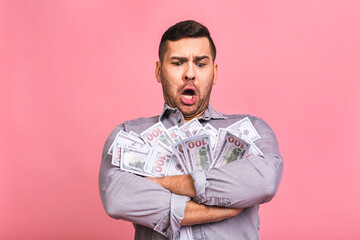 Happy winner! Young rich man in casual holding money dollar bills with surprise isolated over pink background.