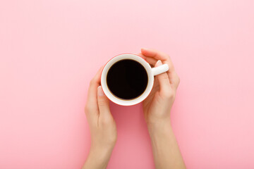 Young adult woman hands holding white cup of black coffee on light pink table background. Pastel color. Closeup. Point of view shot. Top down view.