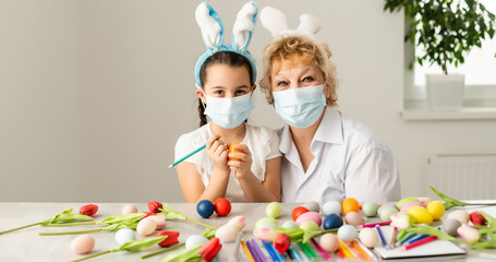 grandmother and granddaughter making easter decoration at home and wearing face mask to protect from coronavirus