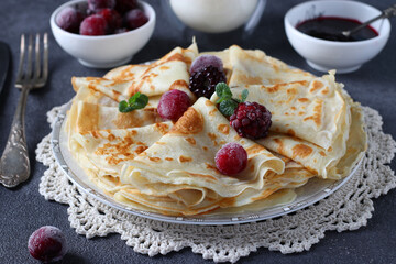 Thin pancakes with wheat flour, eggs and kefir, served with berries, jam and a glass of milk on dark gray table