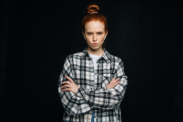 Disgruntled red-haired young woman wearing wireless earphones with crossed hands looking at camera on isolated black background. Pretty redhead lady model emotionally showing facial expressions.