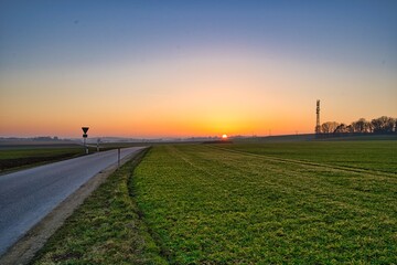 Fototapeta na wymiar Feld und Straße in Bayern, Deutschland mit Blick auf den Sonnenuntergang am Horizont, welcher den Himmel in prächtigen Farben erstrahlen lässt.