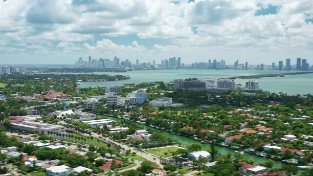 Hyper lapse Miami Beach aerial view of green neighborhood city area with one-story houses. Cityscape on sunny summer day with scenic white clouds in blue sky above green bay waters, 4K drone