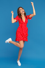 Naklejka premium Successful young woman in red casual dress looking up with excited face with hands up over blue background.