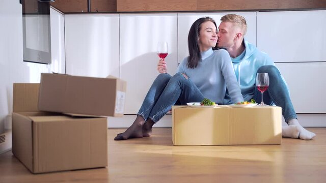 Happy young married couple having lunch and drinking wine on a makeshift table from a cardboard box Cheerful family eating on the floor after moving to a new apartment in home. Newly purchased housing