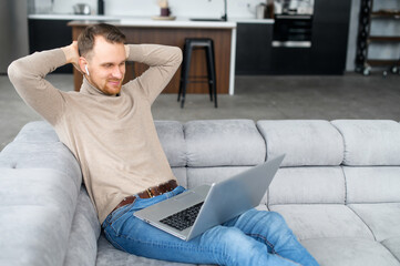 European successful man with earphones sitting on the comfortable pure sofa, smiling, relaxing after productive day with a laptop on the lap, holding hands behind head, happy to end all planned