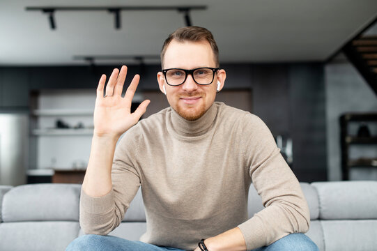 Young Adult European Man In Elegant Black Glasses, Looking At The Camera, Smiling And Waving The Hand, Say Hello, Greeting By Video, Speaking On Webcam, Positive Person, With White Earphones, Stylish