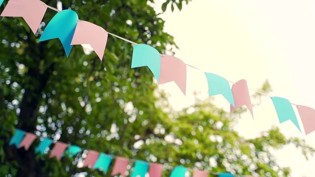 Decoration outside of a birthday celebration. Pastel bunting flags hanging among trees. Summer garden party. Outdoor, wedding. Midsummer, festa junina concept. Natural blurred background, sunny haze.