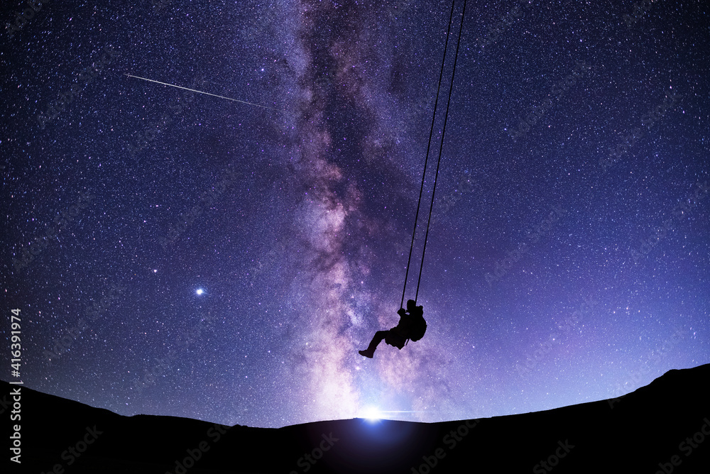 Wall mural fantasy night scene. silhouette of a guy swinging on a swing under bright milky way galaxy. beautifu