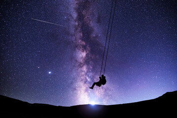 Fantasy night scene. Silhouette of a guy swinging on a swing under bright milky way galaxy. Beautiful starry night. - obrazy, fototapety, plakaty
