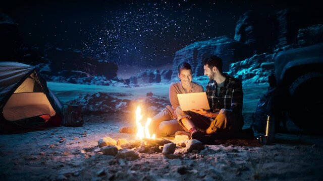 Happy Couple Nature Camping in the Canyon at Night, Use Laptop Computer, Sitting by Campfire. Two Traveling people Have Fun, Smile, Post on Social Media, Watch Funny Video Shows on Streaming Service