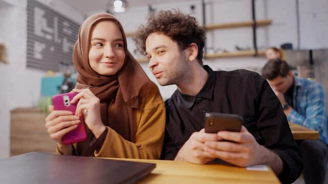 Happy young Muslim couple sitting in cafe using social media messaging on smartphones. Smiling handsome man and beautiful woman surfing Internet and talking in cafeteria. Millennial lifestyle.