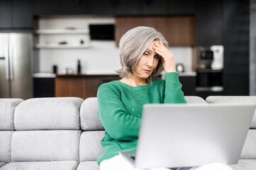 Upset senior woman looks at the laptop screen and feels frustrated, win in lottery, has a problem...