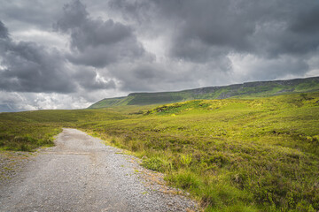 Gravel footpath or trail between green hills and peat bog leading to Cuilcagh Mountain with stormy, dramatic sky in background, Northern Ireland