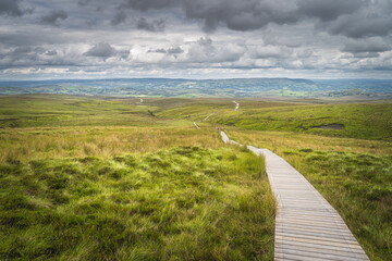 Wooden boardwalk between green hills and bog leading to distant horizon with dramatic sky in...