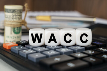 There are cubes on the calculator that say - WACC. Nearby out of focus - dollars, notebook and pen
