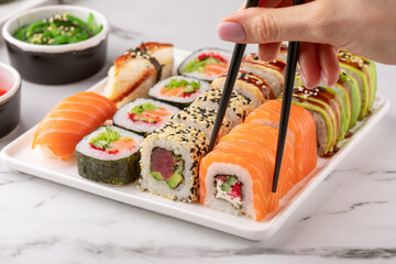 Female hand holds a sushi roll with chopsticks. Sushi rolls set served on a white plate. Traditional Japanese food, angle view.