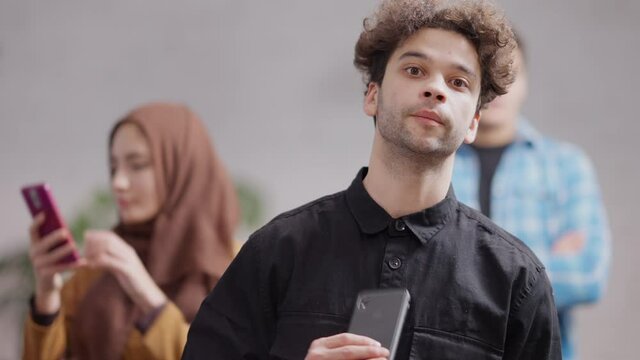Portrait of nervous millennial Middle Eastern man waiting in queue indoors checking time on smartphone. Anxious hurrying young guy in cafeteria or at ATM. City lifestyle.