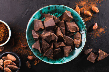 Pieces of dark chocolate in a vintage bowl, cocoa powder and cocoa beans on a dark textured background, top view. Confectionery chocolate background. Chunks of chocolate.
