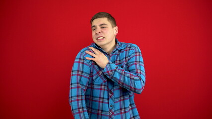 A young man has a sore shoulder and he touches it with his hand. Shooting on a red background.