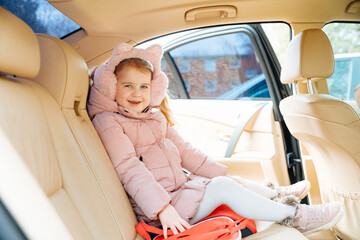 Little blond girl in thick clothes sits in car and smiles. Child on the back seat. Child without a child car seat. Car trip with child. Driving with children in the car in winter