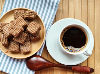 Many square chocolate brown waffles lie on a wooden plate. A cup of black coffee in a white mug. Wooden spoon. Place for text.