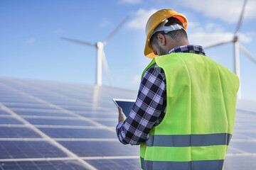 Man working at solar power station with digital tablet - Solar panels with wind turbines in background - Green energy renewable concept