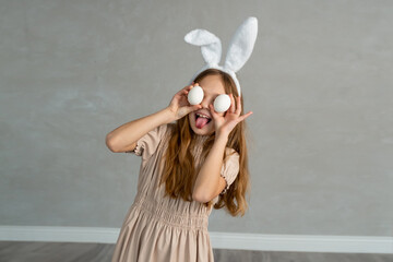 Charming happy Caucasian girl wear bunny ears and holding white easter eggs covering eyes with smiling, shows tongue isolated on gray background. Easter celebration and traditional holiday concept