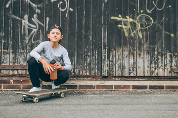 Smiling teenager skateboarder boy sitting beside a wooden grunge graffiti wall with skateboard and...