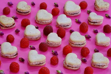 Pink heart homemade macaron cookies filled with raspberry jam for Valentines Day with fresh raspberries and dried roses on a pink background