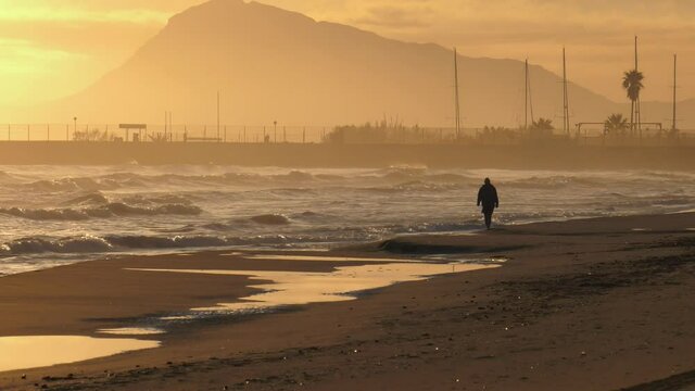Sandy beach at sunrise with old man walking along shore, mountain in background, wide shot. Mediterranean sea, spain
