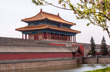 Beijing, China - April 27, 2010: Moat, ramparts, and Red Gate of divine Prowess building as exit of Forbidden City North side under silver cloudscape. Pink flowers.