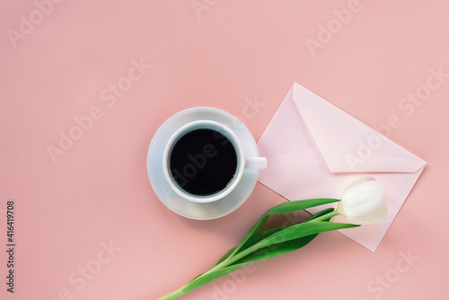 Cup of coffee, envelope and white tulip on coral pink background. Women's day or Mother's day concept. Top view, flat lay.