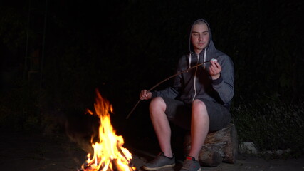 Naklejka premium A young man in a hood fries marshmallows over a fire. Campfire at night.