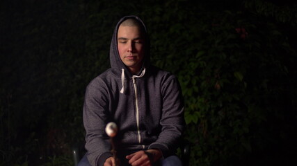 Fototapeta premium A young man in a hood fries marshmallows over a fire and the marshmallows catch fire. Campfire at night.