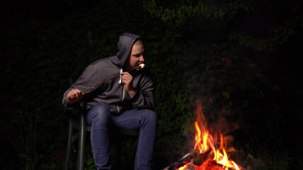 Obraz premium A young man in a hood fries marshmallows over a fire. Campfire at night.