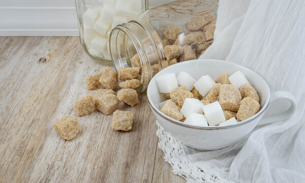 A White Teacup, And Pair Of Open Glass Jars Filled With Sugar Cubes, Raw Sugar Spilling Out
