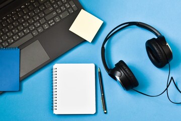 Notepad, earphones with laptop computer on blue background. Podcasting concept. Copy space, flat lay.