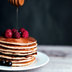 Juicy pancakes with berries and honey on a white plate, spoon, wooden table
